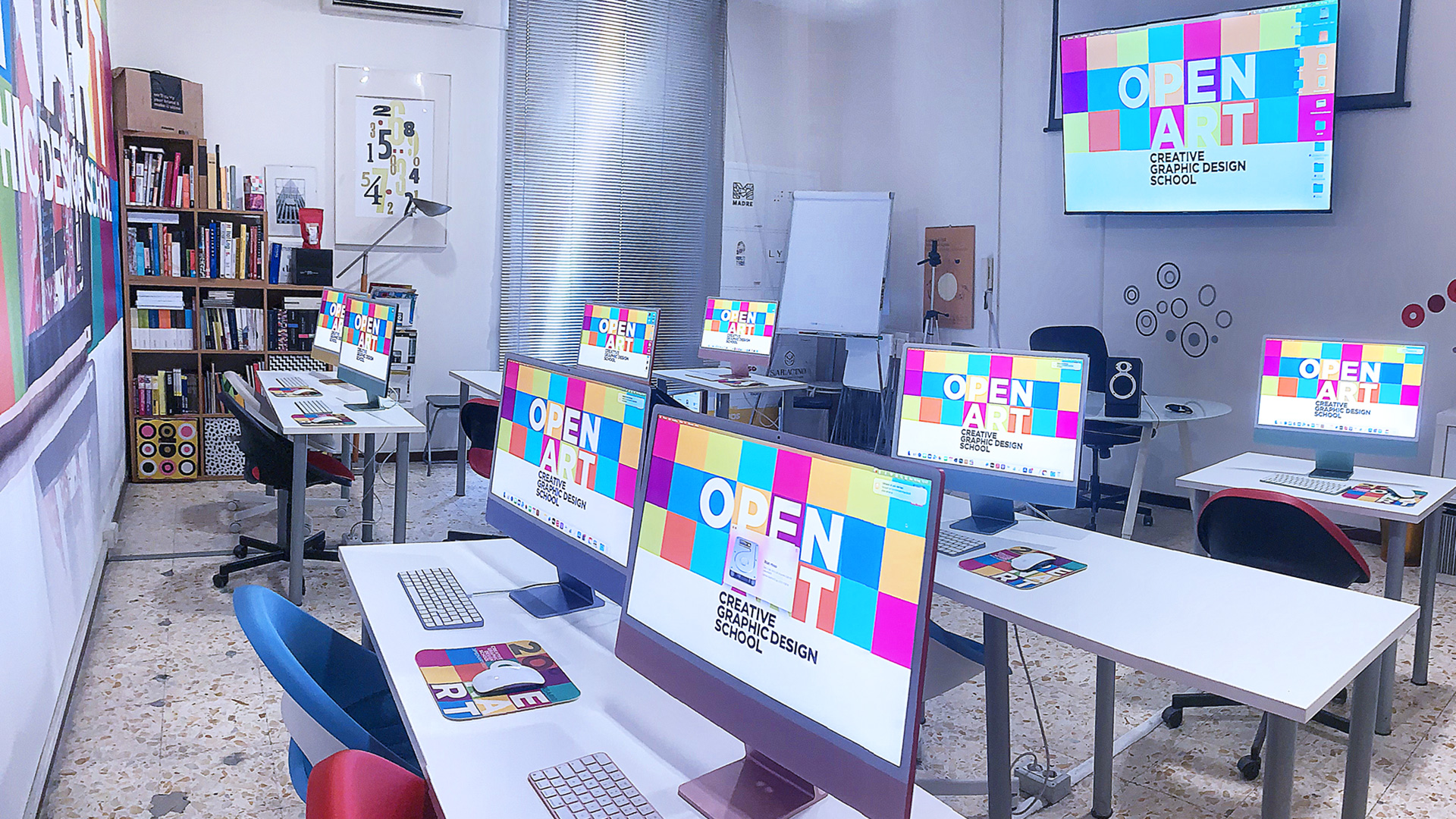 Una classe di dieci studenti in aula con iMac, che lavorano a un progetto di grafica guardando lo schermo del docente