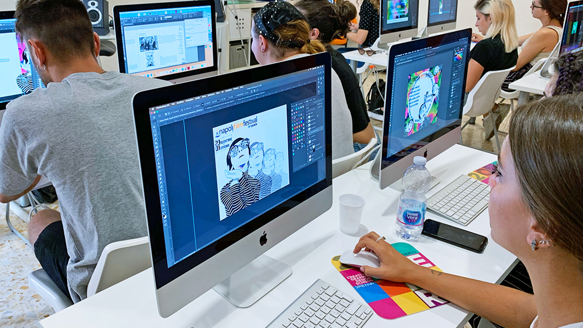 Un'aula luminosa con dieci postazioni iMac, studenti al lavoro concentrati su progetti grafici diversi