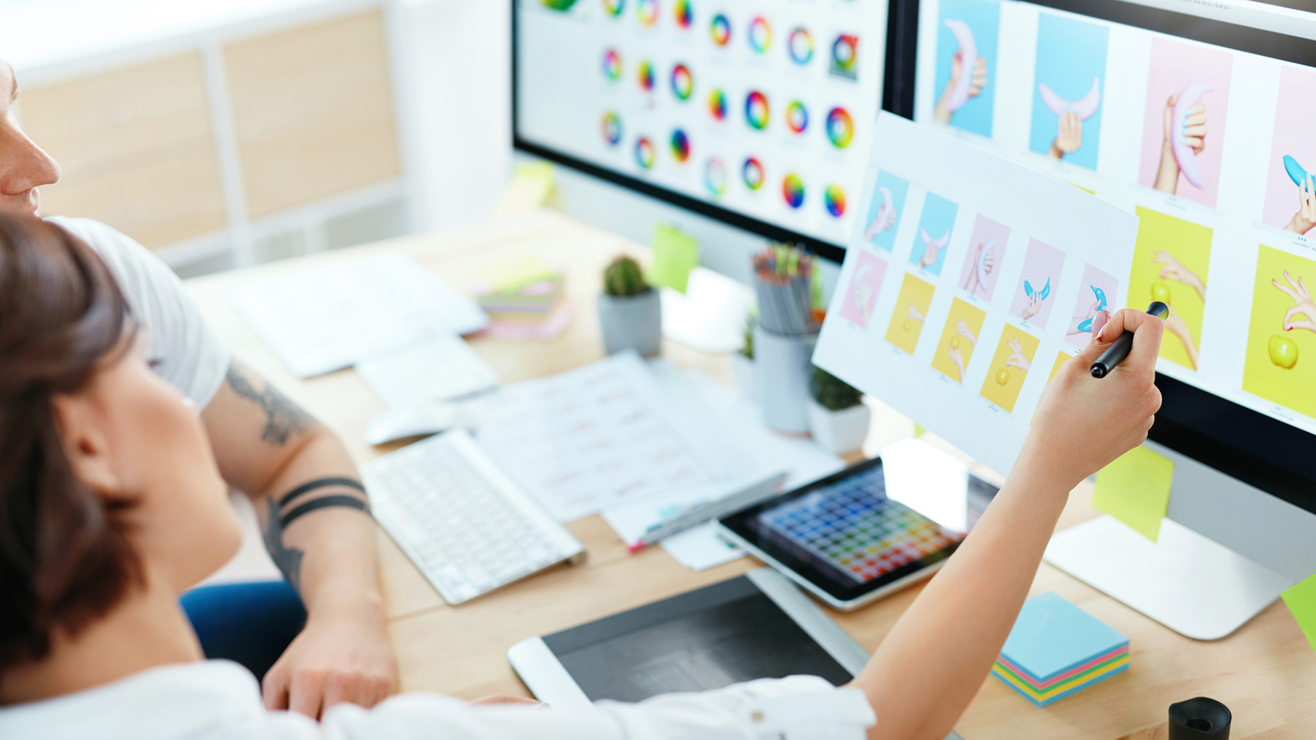 Una studentessa e un docente davanti a un iMac discutono di un progetto di grafica, con altri studenti sullo sfondo